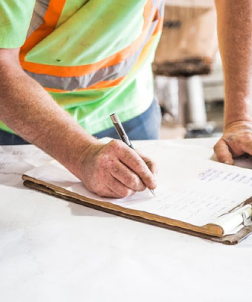 Construction worker wearing a safety vest writing notes on a clipboard placed on a table covered with blueprints.